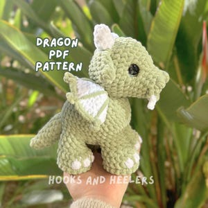 Op de afbeelding: Groen gehaakt draken amigurumi speelgoed met witte vleugels. De draak heeft een schattig gezicht met zwarte ogen en een kleine mond. De tekst "Dragon PDF Pattern" is zichtbaar in de afbeelding.
