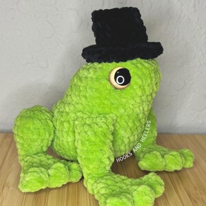 May include: A handmade, plush green frog wearing a black top hat. The frog has large, round eyes and is sitting on a wooden surface. The text "HOOKS AND HEELERS" is visible on the side of the frog.