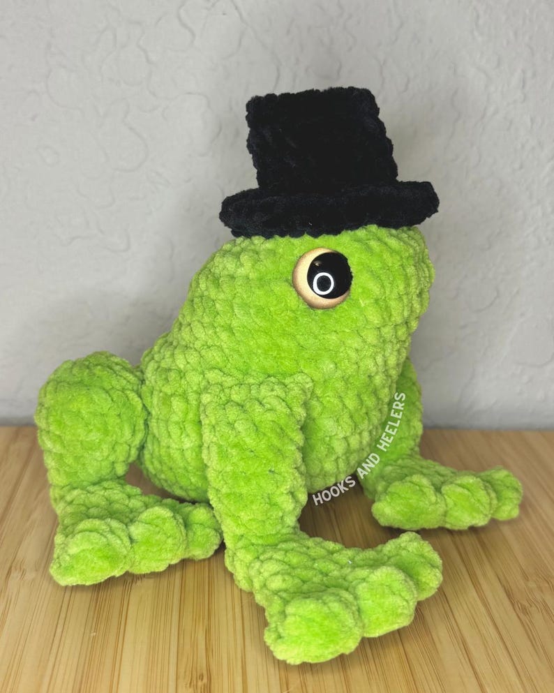 May include: A handmade, plush green frog wearing a black top hat. The frog has large, round eyes and is sitting on a wooden surface. The text "HOOKS AND HEELERS" is visible on the side of the frog.
