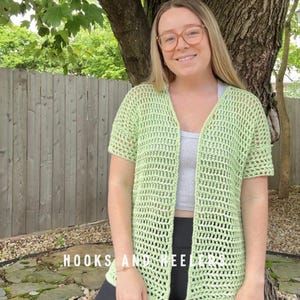 May include: A light green, open-front crochet cardigan with short sleeves. The cardigan is made of a loosely woven material. The text "HOOKS AND NEEDLES" is visible at the bottom of the image. The cardigan is worn over a gray top.