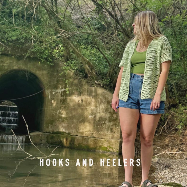 May include: A person wearing a light green crochet cardigan over a green tank top and denim shorts. The text "HOOKS AND HEELERS" is at the bottom of the image. The background includes a tunnel and trees.