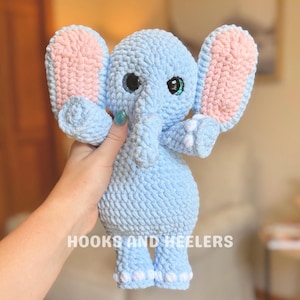 May include: A light blue crochet elephant with pink inner ears and large, sparkly eyes. The handmade stuffed animal has a trunk, small feet, and the text "HOOKS AND HEELERS" at the bottom. A cute and cuddly toy.