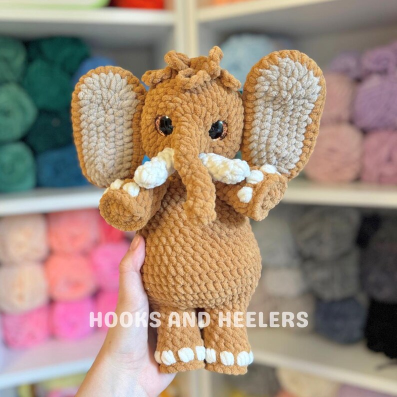 May include: A handmade, plush elephant toy in shades of brown and beige. The elephant has large, floppy ears, white tusks, and small, black eyes. The text "HOOKS AND HEELERS" is printed at the bottom. The toy is soft and cuddly.