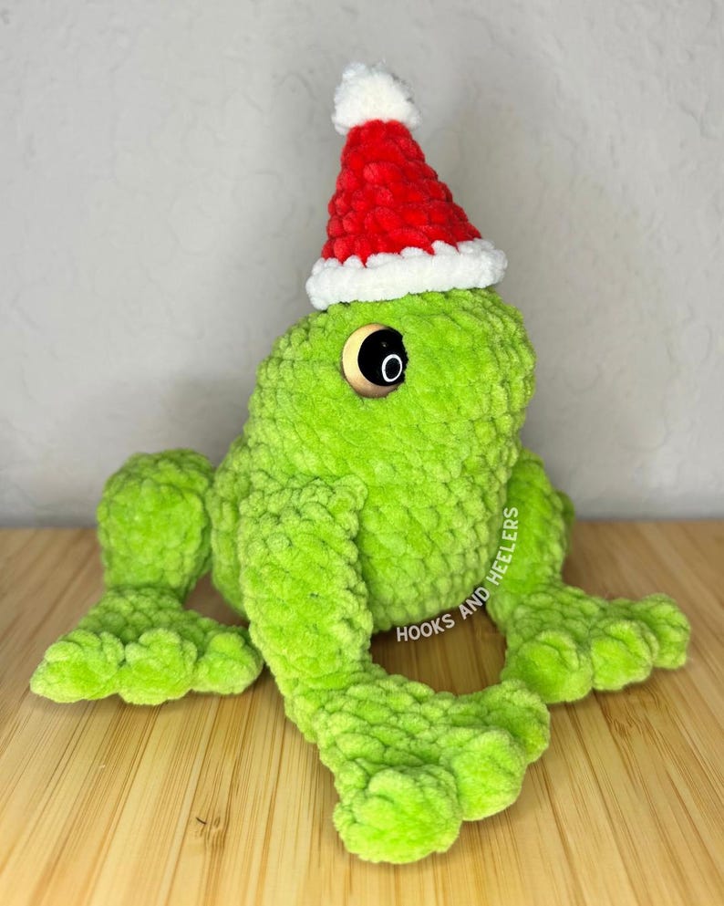 May include: A plush green frog wearing a red and white Santa hat. The frog has large black eyes and is sitting on a wooden surface. The text "HOOKS AND HEELERS" is visible on the frog's leg. A festive, handmade item.