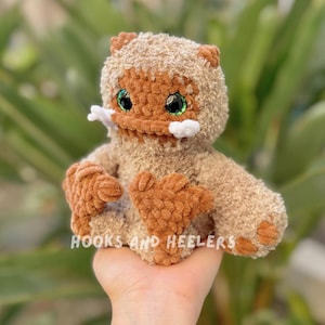 May include: A crocheted plush toy with brown fur, green eyes, and white teeth. The toy is sitting in a person's hand.