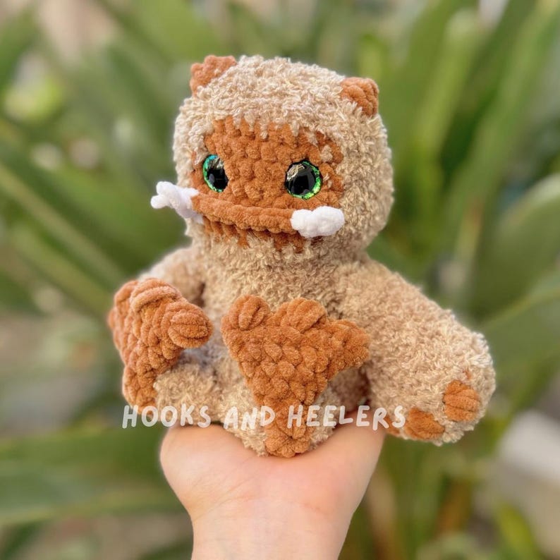 May include: A crocheted plush toy with brown fur, green eyes, and white teeth. The toy is sitting in a person's hand.