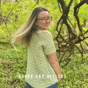 May include: A light green, open-weave crochet top with short sleeves. The top is made of a soft, textured yarn and has a relaxed fit. The text "HOOKS AND HEELERS" is visible at the bottom of the image.