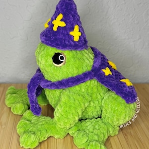 May include: A handmade, plush green frog wearing a purple wizard hat and scarf, adorned with yellow stars. The frog has large, round eyes and is sitting on a wooden surface. The hat and scarf add a whimsical touch to the soft, textured toy.