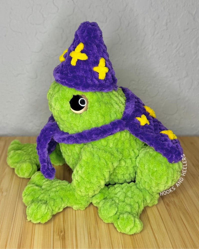 May include: A handmade, plush green frog wearing a purple wizard hat and scarf, adorned with yellow stars. The frog has large, round eyes and is sitting on a wooden surface. The hat and scarf add a whimsical touch to the soft, textured toy.