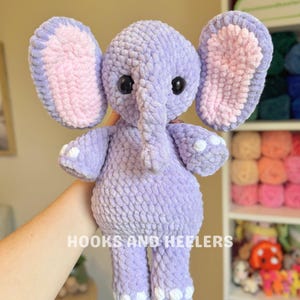 May include: A handmade, lavender crochet elephant with large ears, a trunk, and small white accents on its feet. The inside of the ears is a soft pink. The elephant has large black eyes. The text "HOOKS AND HEELERS" is visible.