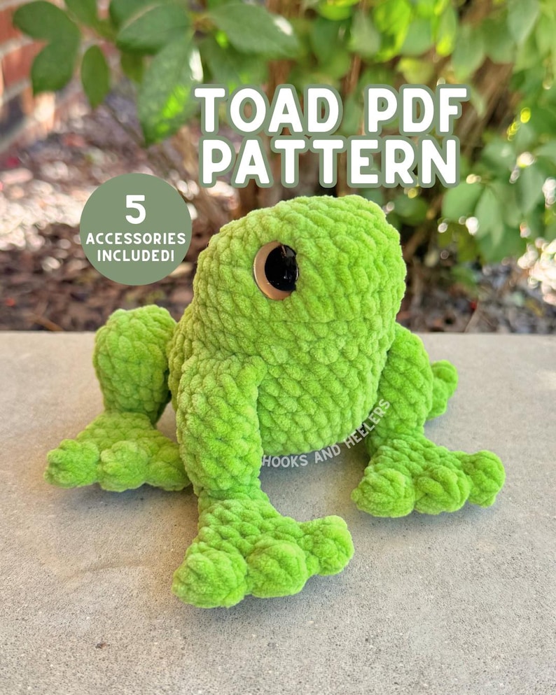 May include: A plush green toad with large, round eyes and a textured surface. The image includes the text "TOAD PDF PATTERN" and "5 ACCESSORIES INCLUDED!" The toad is positioned on a gray surface with a blurred green background.