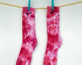 Puede incluir: Dos pares de calcetines teñidos con tinte anudado en tonos rosa y blanco, colgados de una cuerda amarilla con pinzas azules. Cada calcetín tiene un logotipo Nike negro. Accesorio colorido.