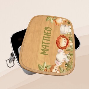 Könnte beinhalten: Eine braune Holzlunchbox mit einem Klappdeckel. Der Deckel hat ein farbenfrohes Design mit Dschungeltieren und den Namen "MATTHEW" in grünen Buchstaben.