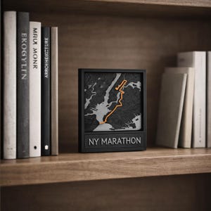 Op de afbeelding: Een zwart omkaderde kaart van de NY Marathon-route, met een oranje lijn die de route markeert, tegen een grijze en zwarte achtergrond. De tekst "NY MARATHON" wordt onder de kaart weergegeven. Het item wordt tentoongesteld op een houten plank met boeken.