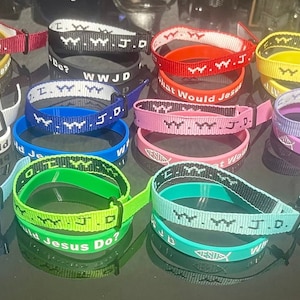 May include: A collection of colorful wristbands with the text "WWJD" (What Would Jesus Do?) and a fish symbol. The wristbands are made of woven fabric and come in a variety of colors, including red, yellow, blue, green, and purple.