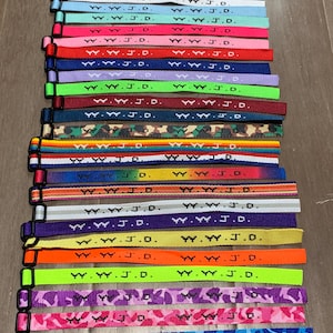 Könnte beinhalten: Eine Sammlung von 18 bunten Armbändern mit dem aufgedruckten Text "W.W.J.D.". Die Armbänder haben verschiedene Muster, darunter Streifen, Tarnmuster und einfarbige Farben.