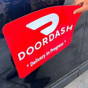 May include: A red DoorDash magnet with a white logo and the text "Delivery in Progress" on a black car.