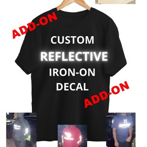 May include: Black t-shirt with the text "CUSTOM REFLECTIVE IRON-ON DECAL" in white. The text "ADD-ON" is written in red on the top left and bottom right corners of the shirt. The shirt is hanging on a wooden hanger. Three images of people wearing the shirt with different reflective designs are shown below.