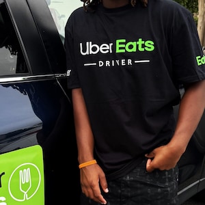 Peut inclure: T-shirt noir avec le logo Uber Eats et le mot « DRIVER » en blanc et vert. Un autocollant de livraison Uber Eats vert et blanc est sur le côté d'une voiture noire. La personne porte un pantalon noir à motifs et un bracelet orange.
