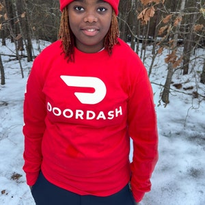 Op de afbeelding: Een persoon draagt een rood shirt met lange mouwen met het DoorDash-logo op de voorkant en een rode beanie met het DoorDash-logo op de voorkant. De persoon staat in een besneeuwd bos.