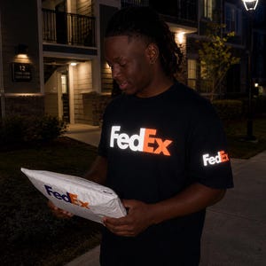 以下が含まれることがあります： 胸元と左袖に白とオレンジのFedExロゴが入った黒の半袖Tシャツ。太ももに小さな白いグラフィックが入った黒いパンツと組み合わされています。白いFedExの配送用封筒が手に持たれています。
