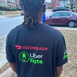 May include: A person wearing a black t-shirt with the logos for DoorDash and Uber Eats. The logos are in red, white, and green.
