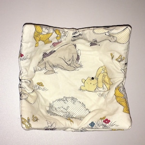 May include: A white fabric square with a Winnie the Pooh theme. The fabric features a yellow, brown, and black print of Winnie the Pooh, Piglet, Eeyore, and Tigger.