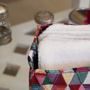 Peut inclure: Une boîte de rangement en tissu colorée avec un motif géométrique de triangles dans des tons de rouge, jaune, vert, bleu et blanc. La boîte est remplie de débarbouillettes en coton blanc.