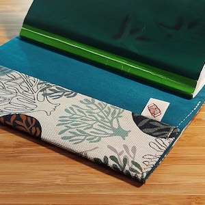 May include: A fabric notebook cover featuring a white base with a teal and dark blue coral reef pattern. The interior is lined with solid teal fabric and includes a small white tag with a red spool icon. A green notebook is inserted inside.