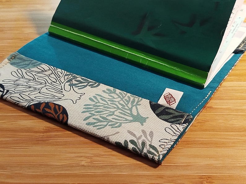 May include: A fabric notebook cover featuring a white base with a teal and dark blue coral reef pattern. The interior is lined with solid teal fabric and includes a small white tag with a red spool icon. A green notebook is inserted inside.