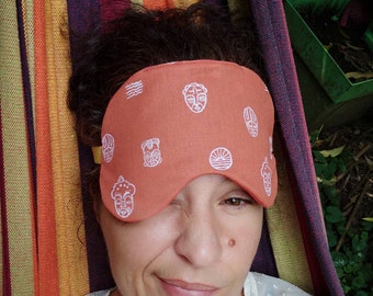 May include: A terracotta-colored sleep mask featuring a repeating white line-art pattern of stylized tribal masks and sun icons. The fabric mask is contoured to fit over the eyes and is secured with a yellow elastic strap.