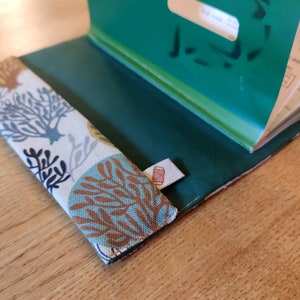 May include: A fabric book cover featuring a white background with a repeating pattern of blue, teal, and brown coral-like shapes. The interior is a solid dark green fabric, and a small white tag with a red spool icon is attached to the side.