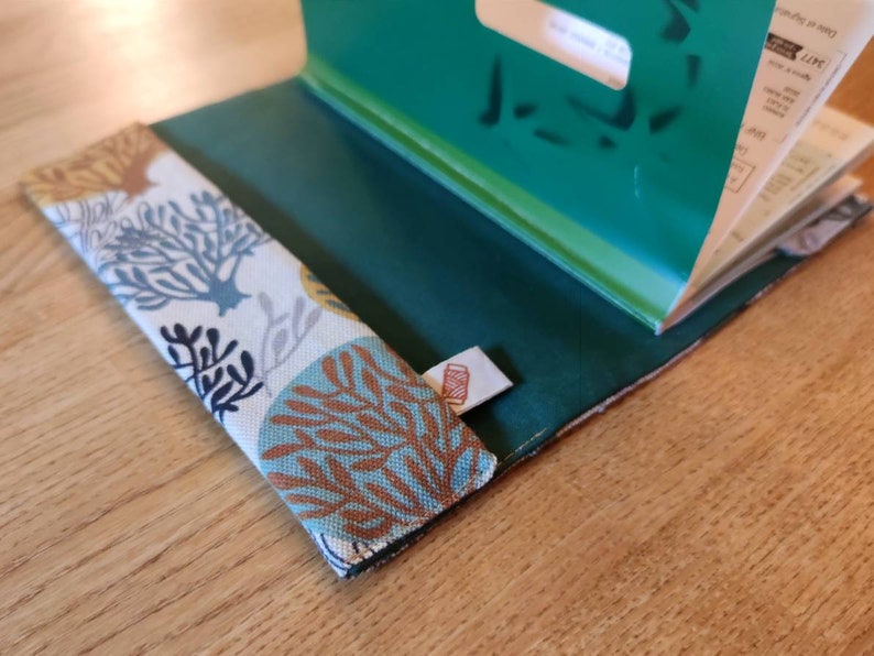 May include: A fabric book cover featuring a white background with a repeating pattern of blue, teal, and brown coral-like shapes. The interior is a solid dark green fabric, and a small white tag with a red spool icon is attached to the side.