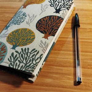 May include: A rectangular notebook with a fabric cover featuring a repeating pattern of stylized trees in shades of mustard yellow, dark teal, and brown on an off-white background, positioned next to a clear plastic ballpoint pen with black ink.