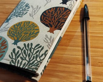 May include: A rectangular notebook with a fabric cover featuring a repeating pattern of stylized trees in shades of mustard yellow, dark teal, and brown on an off-white background, positioned next to a clear plastic ballpoint pen with black ink.