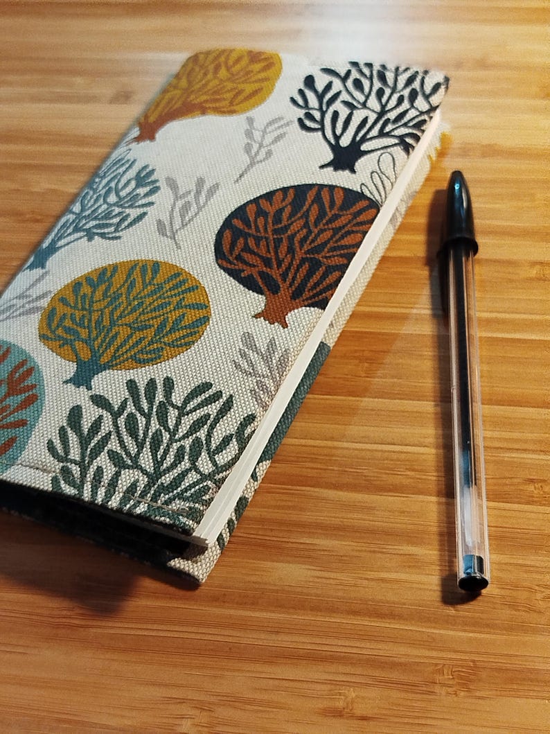 May include: A rectangular notebook with a fabric cover featuring a repeating pattern of stylized trees in shades of mustard yellow, dark teal, and brown on an off-white background, positioned next to a clear plastic ballpoint pen with black ink.
