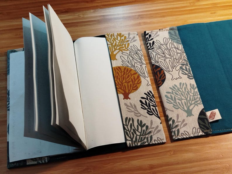 May include: A fabric notebook cover featuring a cream-colored exterior with a repeating pattern of stylized coral and trees in shades of mustard yellow, teal, and black, paired with a solid teal interior lining and a small white brand tag.