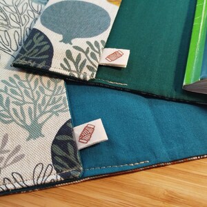 May include: Two fabric book covers featuring a cream-colored exterior with a repeating pattern of stylized trees and circular shapes in shades of blue, green, and brown. The interior is lined with solid teal fabric and includes a small white tag with a red spool icon.