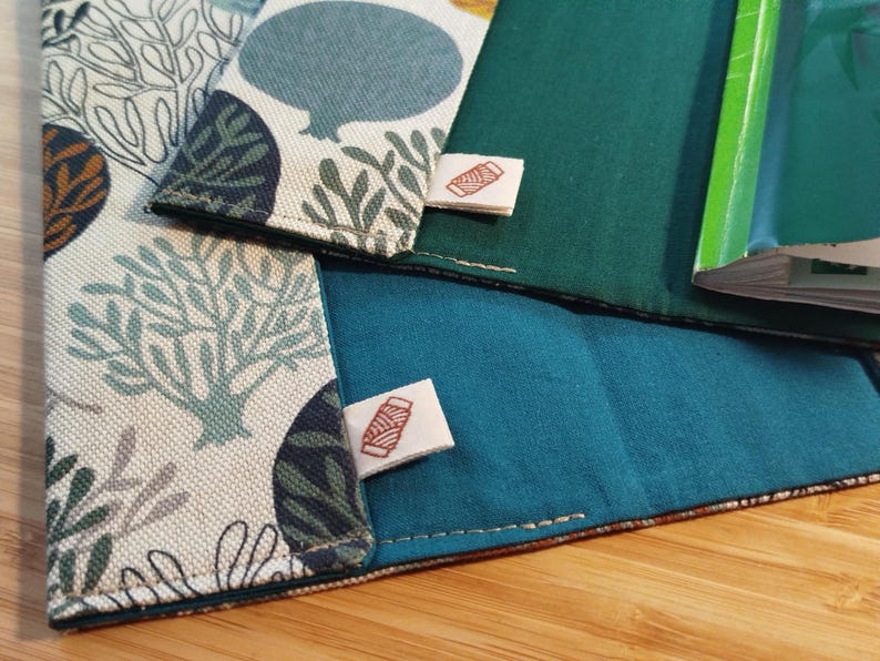 May include: Two fabric book covers featuring a cream-colored exterior with a repeating pattern of stylized trees and circular shapes in shades of blue, green, and brown. The interior is lined with solid teal fabric and includes a small white tag with a red spool icon.