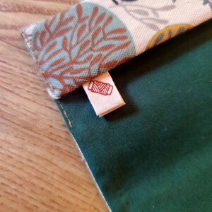 May include: A fabric pouch featuring a solid dark green section and a folded top flap with a cream background patterned with teal and rust-colored coral-like designs. A small white fabric tag with a red spool of thread icon is attached.