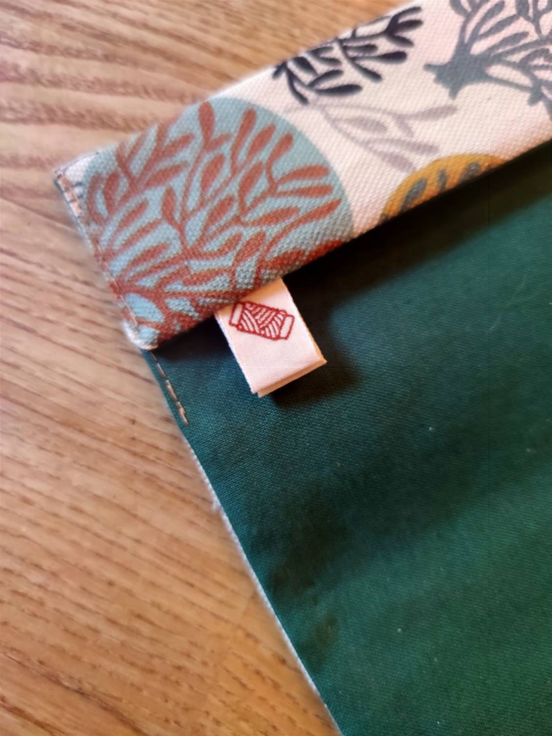 May include: A fabric pouch featuring a solid dark green section and a folded top flap with a cream background patterned with teal and rust-colored coral-like designs. A small white fabric tag with a red spool of thread icon is attached.