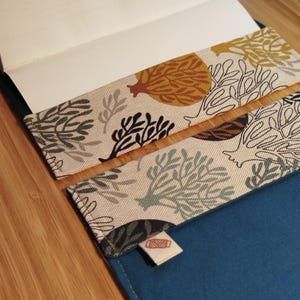 May include: A fabric book cover featuring a beige background with a repeating pattern of stylized trees and coral in shades of mustard yellow, dark green, and black. The interior is solid blue with a small white tag showing a red thread spool icon.