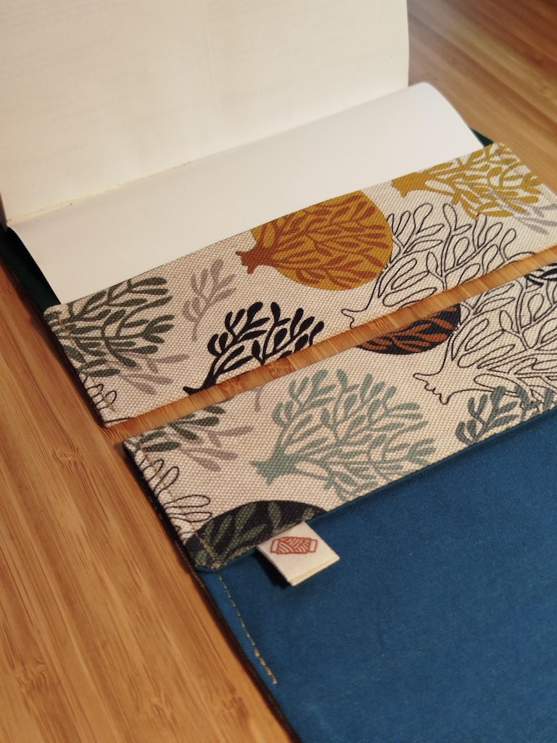 May include: A fabric book cover featuring a beige background with a repeating pattern of stylized trees and coral in shades of mustard yellow, dark green, and black. The interior is solid blue with a small white tag showing a red thread spool icon.