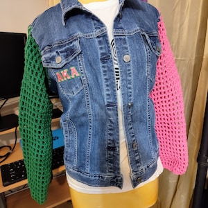 May include: A blue denim jacket with green and pink crocheted sleeves. The jacket has a patch on the left breast that says "AKA" in pink letters.