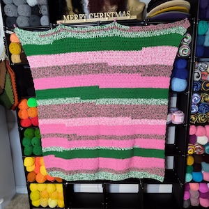 Op de afbeelding: Een handgemaakte gehaakte deken in roze, groene en grijze tinten. De deken heeft horizontale strepen van verschillende breedtes. De tekst "MERRY CHRISTMAS" is zichtbaar boven de deken. De deken wordt tentoongesteld op een zwart rek.