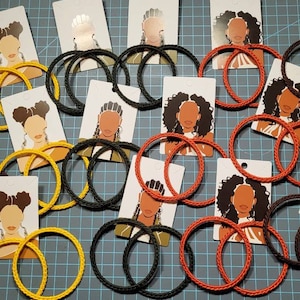 May include: A collection of handmade hoop earrings in yellow, orange, and black. The earrings are made of braided cord and are displayed on cards with illustrations of people wearing the earrings.