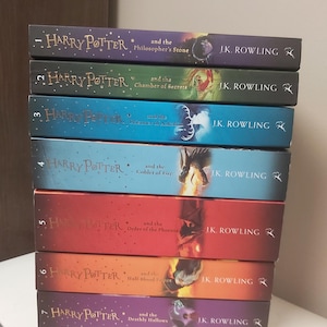 Può includere: Una pila di sette libri di Harry Potter di J.K. Rowling. I dorsi dei libri sono di vari colori, tra cui viola, rosso, blu e verde. Ogni titolo del libro è visibile, con il nome dell'autore su ogni dorso.
