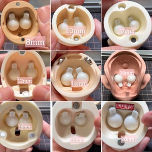 Puede incluir: Nueve imágenes de primer plano de cuencas de ojos de muñeca con insertos de ojos de diferentes tamaños. Los insertos de ojos son blancos y tienen un pequeño pomo en la parte superior. Las cuencas están etiquetadas con el tamaño del inserto de ojos en milímetros: 8 mm, 10 mm, 12 mm, 14 mm, 16 mm, 18 mm y 20 mm.