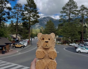 May include: A light brown wooden bear figurine featuring carved fur textures, two dark circular eyes, and a simple carved nose and mouth. The bear is depicted in a seated position with visible chisel marks throughout the surface of the wood.