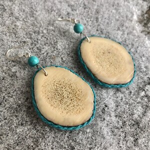 May include: A pair of handmade earrings featuring oval-shaped bone pendants with a turquoise blue rope trim. Each earring has a silver hook and a small turquoise bead. The earrings are displayed on a textured gray surface.